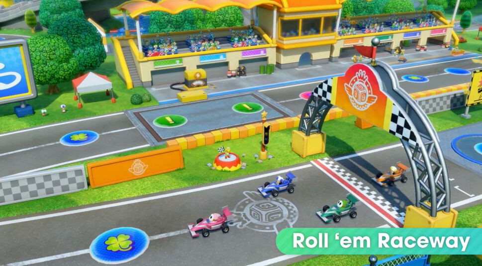 Auf diesem Bild aus dem Super Mario Party-Jamboree-Test sieht man die "Roll 'em Raceway", eine farbenfrohe Rennstrecke mit kleinen Formel-1-ähnlichen Autos. Drei Rennautos in den Farben Pink, Blau und Grün fahren auf der Strecke über die Ziellinie, während Zuschauer in der Tribüne im Hintergrund sitzen. Ein großes Zielflaggen-Tor markiert den Zielbereich, und der Bereich um die Strecke ist lebendig gestaltet mit Bäumen, Tribünen und Renn-Elementen. Die Atmosphäre ist fröhlich und dynamisch, passend zu einer bunten Mario Party-Umgebung.