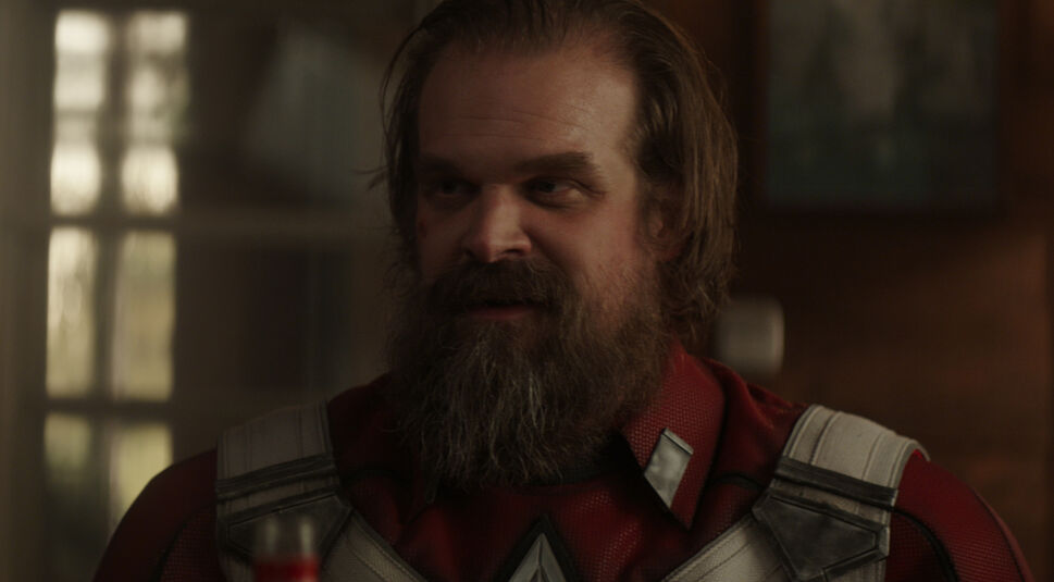 Auf dem Bild vom Thunderbolts-Team-Artikel ist Alexei Shostakov, auch bekannt als Red Guardian, aus dem Film Black Widow zu sehen. Er trägt sein rotes Kostüm mit markanten weißen und grauen Akzenten, das an seine Vergangenheit als russischer Superheld erinnert. Alexei hat einen dichten Bart und lange Haare, die ihm ein robustes, leicht verwittertes Aussehen verleihen. Er befindet sich in einer gemütlich beleuchteten Innenraumszene, die eine entspannte, aber nachdenkliche Stimmung erzeugt. Sein Gesichtsausdruck wirkt zufrieden und leicht verschmitzt, als wäre er in ein vertrautes Gespräch vertieft.