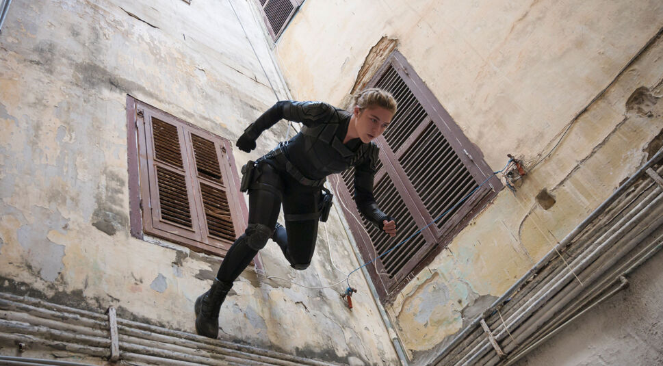 Auf dem Bild vom Thunderbolts-Team-Artikel ist Yelena Belova, bekannt aus dem Film Black Widow, in einer intensiven Action-Sequenz zu sehen. Sie trägt ein schwarzes taktisches Outfit mit Schutzpolstern und Ausrüstungsgürtel, während sie sich mitten im Sprung befindet. Der Schauplatz scheint eine enge, etwas heruntergekommene Hinterhofgasse zu sein, mit alten, abblätternden Wänden und rostigen Fensterläden. Die Szene vermittelt eine dynamische, spannende Atmosphäre, typisch für einen Spionage- oder Action-Thriller.