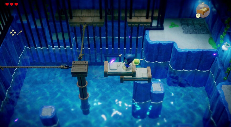 Auf dem Bild zu "Zelda: Echoes of Wisdom Tipps" ist eine Figur mit blauer Kleidung und gelbem Schild in einer rätselhaften Höhlenumgebung zu sehen. Die Figur steht auf einer kleinen Plattform über klarem, blauem Wasser, umgeben von Gitterstäben und Holzbrücken. Links oben sind Herzen und Symbole für den Spielfortschritt erkennbar. Die Szene deutet auf ein Rätsel hin, das gelöst werden muss, um weiterzukommen.