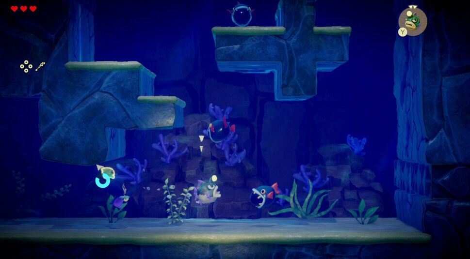Auf dem Bild zu "Zelda: Echoes of Wisdom Tipps" befindet sich Zelda in einer Unterwasserwelt. Umgeben von Fischen und Korallen navigiert sie durch felsige Strukturen, während Gegner in Form von großen, kugelförmigen Fischen mit roten Flossen auftauchen. Die blaue Unterwasseratmosphäre wird durch schimmernde Lichteffekte und Pflanzen verstärkt. Oben links zeigt die Anzeige ihre verbleibenden Herzen und Fortschrittssymbole.