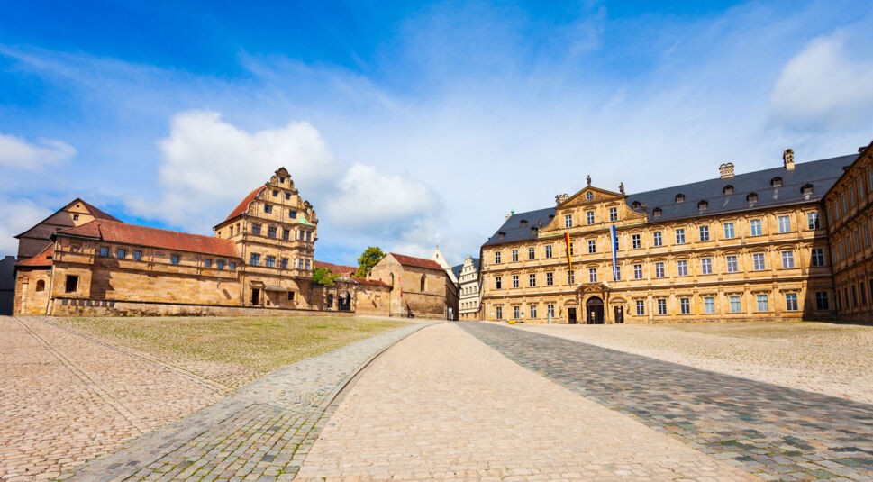 Das Bild zeigt den Domplatz in Bamberg, einen der Drehorte der Serie "Die Kaiserin". Der Platz ist umgeben von historischen Gebäuden, darunter das Alte Hofhaltung und das barocke Neue Residenz-Gebäude. Das Kopfsteinpflaster und der blaue Himmel schaffen eine harmonische und historische Kulisse.