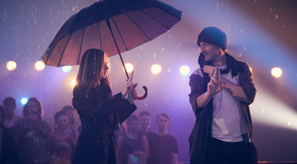 Auf dem Bild in dem Artikel über Robbie Williams' Film Better Man, sieht man den Regisseur Michael Gracey mit einer Frau, die unter einem Regenschirm steht. Im Hintergrund stehen weitere Menschen und es leuchten Lichter.