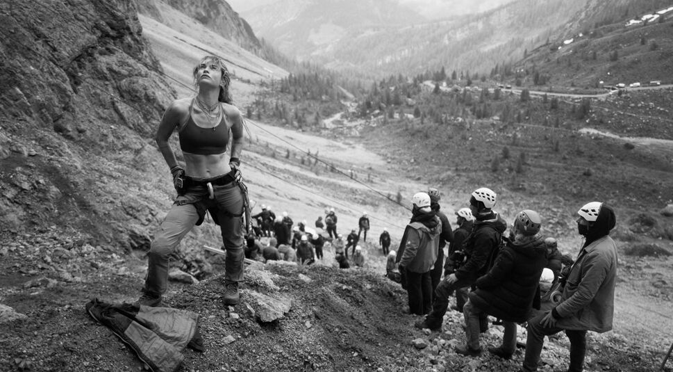 Auf dem Bild in dem Artikel zum Cliffhanger-Reboot ist Lily James am Set zu sehen. Sie steht auf einem Berg und im Hintergrund sind weitere Menschen zu sehen. Das Bild ist schwarz-weiß.