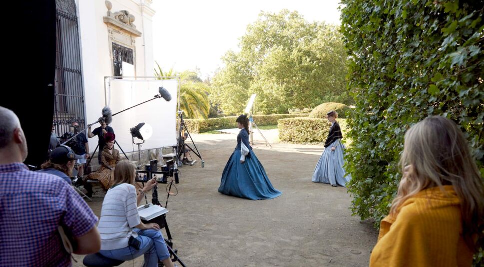 Das Bild zeigt eine Hinter-den-Kulissen-Szene der Serie "Die Kaiserin" an einem der Drehorte. Zwei Schauspielerinnen in aufwendigen historischen Kleidern bewegen sich durch einen Garten, während die Crew mit Kameras, Mikrofonen und Beleuchtung arbeitet. Im Vordergrund koordinieren Mitarbeiter die Szene, während Sonnenschein und grüne Büsche die elegante Kulisse unterstreichen.