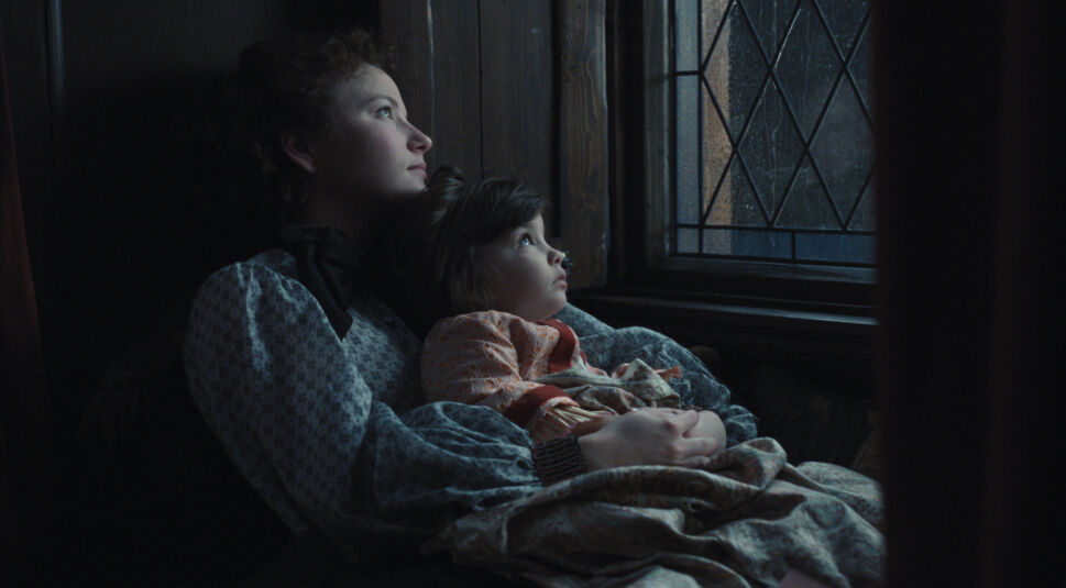 Auf dem Bild zur Serie "Die Kaiserin Staffel 2 Ende erklärt" sind Devrim Lingnau als Kaiserin Elisabeth, auch Sisi genannt, und ihre Tochter zu sehen. Beide sitzen eng aneinander gekuschelt an einem Fenster mit verzierten Glasmustern und blicken gedankenverloren nach draußen. Sisi trägt eine dezente, gemusterte Bluse mit einer schwarzen Schleife, während ihre Tochter in ein farbenfrohes Kleid gehüllt ist. Die dunkle und ruhige Umgebung, ergänzt durch das weiche Licht, erzeugt eine melancholische und nachdenkliche Atmosphäre, die auf eine emotionale Schlüsselszene hinweist.