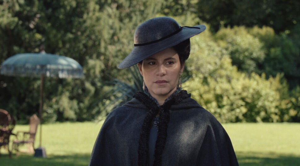 Auf dem Bild zur Serie "Die Kaiserin Staffel 2 Ende erklärt" ist Almila Bagriacik als Leontine von Apafi zu sehen. Sie trägt einen eleganten, dunklen Umhang und einen breitkrempigen schwarzen Hut, die ihre geheimnisvolle Ausstrahlung betonen. Ihre ernste Miene deutet auf eine angespannte oder bedeutende Szene hin. Im Hintergrund ist ein sonniger Garten mit viel Grün sowie ein antiker Sonnenschirm und Gartenmöbel zu erkennen, die eine kontrastreiche, ruhige Atmosphäre schaffen.