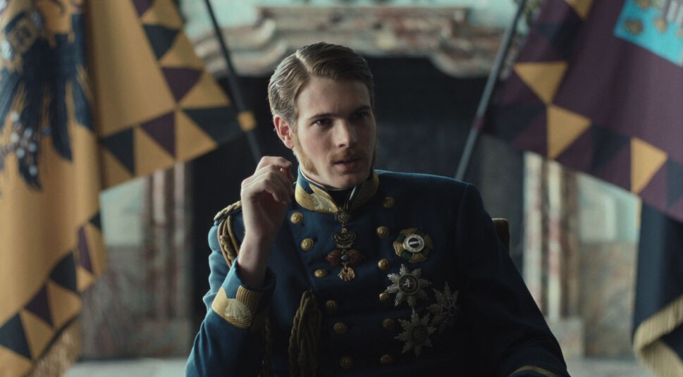 Auf dem Bild zur Serie "Die Kaiserin Staffel 2 Ende erklärt" ist Philip Froissant als Kaiser Franz zu sehen. Er trägt eine reich verzierte, dunkelblaue Uniform mit zahlreichen Orden und Abzeichen, die seinen Rang betonen. Im Hintergrund sind kaiserliche Fahnen in Gelb und Schwarz zu erkennen, die die majestätische und historische Atmosphäre unterstreichen. Der Charakter wirkt nachdenklich und konzentriert, was auf eine wichtige Entscheidungsszene hindeuten könnte.