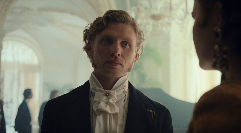 Auf dem Bild zur Serie "Die Kaiserin Staffel 2 Ende erklärt" ist Johannes Nussbaum als Erzherzog Maximilian zu sehen. Er trägt ein elegantes dunkles Jackett mit einem weißen Halstuch, das seinen aristokratischen Status unterstreicht. An seiner Jacke ist eine goldene Brosche in Form einer Libelle befestigt. Im Hintergrund ist ein opulent dekorierter Raum mit Kronleuchtern und weiteren Personen in formeller Kleidung zu erkennen. Maximilian blickt nachdenklich zur Seite, während ein Gespräch im Vordergrund angedeutet wird, was auf eine spannende Wendung hinweist.