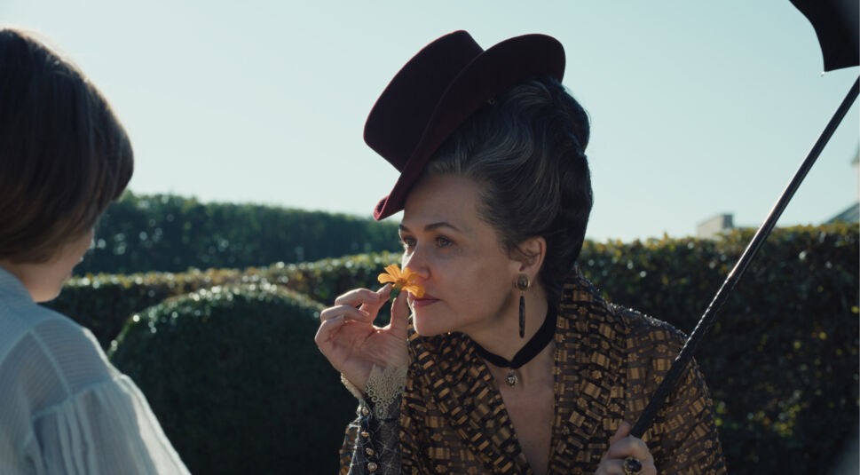 Auf dem Bild zur Serie "Die Kaiserin Staffel 2 Ende erklärt" ist Melika Foroutan als Erzherzogin Sophie zu sehen. Sie trägt ein elegantes, gemustertes Kleid und einen auffälligen, dunkelroten Hut. In der Hand hält sie eine gelbe Blume, die sie nachdenklich betrachtet und daran riecht. Ihre Haltung und der intensive Blick vermitteln einen Moment der Reflexion. Der sonnige Garten im Hintergrund mit geschnittenen Hecken schafft eine friedliche, aber zugleich angespannte Atmosphäre.