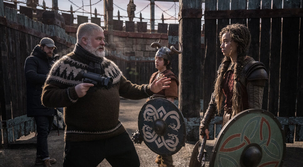 Auf diesem Bild vom Set zu "Drachenzähmen leicht gemacht 2025" ist Regisseur Dean DeBlois links zu sehen, wie er mit einer Kameraanweisung gestikuliert. Rechts steht Nico Parker in einem Wikingerkostüm mit einem grünen Schild und einer Axt. Im Hintergrund ist eine historische Szenerie mit Holzzäunen und weiteren Crew-Mitgliedern zu erkennen. Die Atmosphäre wirkt konzentriert und lebendig, passend zu den Vorbereitungen einer Filmszene.