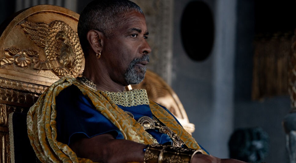 Dieses Bild aus Gladiator 2 zeigt Macrinus, gespielt von Denzel Washington, in einer majestätischen Pose. Er sitzt ernst und nachdenklich auf einem reich verzierten Thron mit goldenen Details, die seine Macht und Autorität unterstreichen. Gekleidet in eine königliche, dunkelblaue Robe mit goldenen Verzierungen und einem Umhang, strahlt er eine würdige und respektvolle Präsenz aus. Sein konzentrierter Blick vermittelt Entschlossenheit und Tiefe und deutet auf seine bedeutende Rolle innerhalb der Handlung hin.