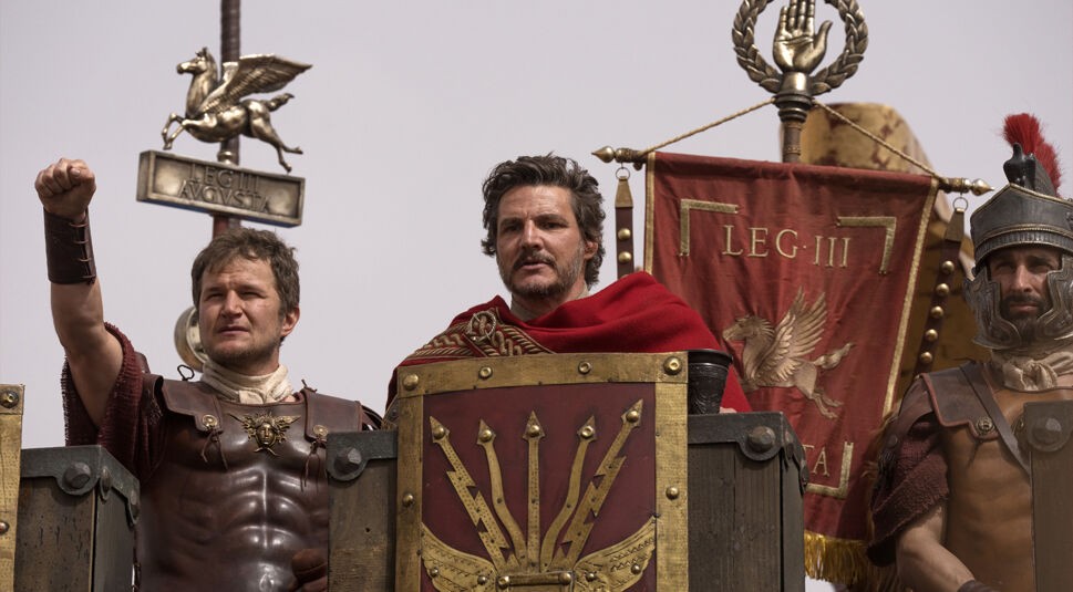 In dieser Szene aus Gladiator 2 ist Marcus Acacius, gespielt von Pedro Pascal, in römischer Rüstung zu sehen. Er steht ernst und entschlossen, umgeben von weiteren Soldaten und Bannern der Legion III. Links von ihm hebt ein Legionär entschlossen die Faust, während im Hintergrund Standarten und Symbole der römischen Legionen hervorragen. Die Atmosphäre ist feierlich und von militärischer Stärke geprägt.