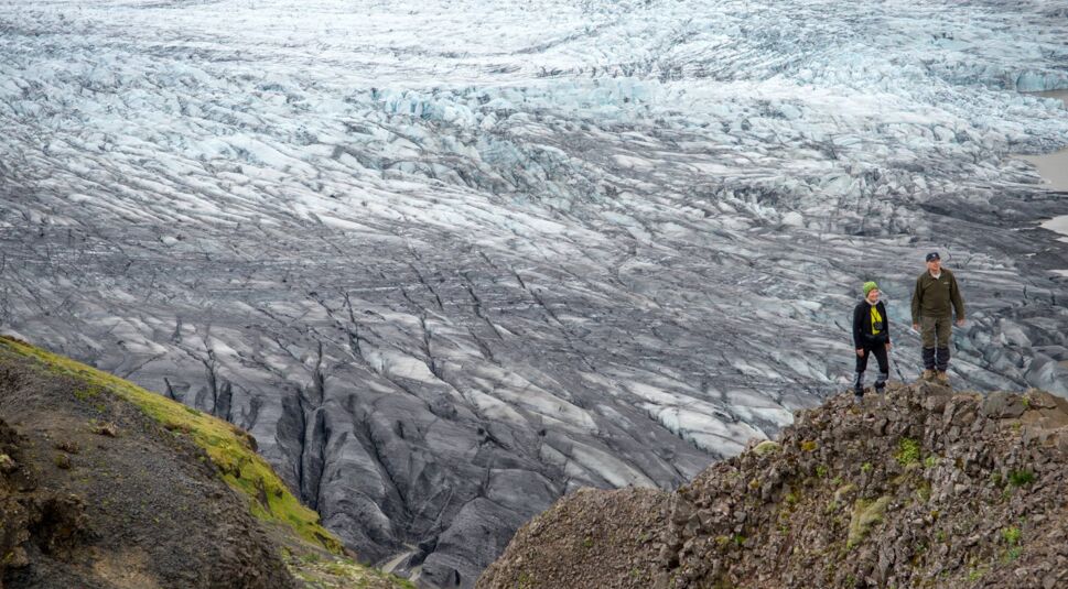 Auf diesem Bild zu Interstellar Fakten ist der isländische Gletscher Svínafellsjökull zu sehen, der als Kulisse für Planetenaufnahmen im Film Interstellar diente. Zwei Personen stehen auf einem felsigen Vorsprung und betrachten die eindrucksvolle, eisige Landschaft des Gletschers, die mit ihren grauen und bläulichen Tönen eine raue, außerirdische Atmosphäre vermittelt.