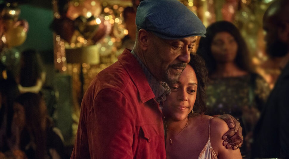 Auf dem Bild zur Serie "Ich vermisse Dich" sind Lenny Henry als Clint Donovan und Rosalind Elezar als Kat Donovan zu sehen. Clint trägt eine blaue Mütze und eine rote Jacke und hält Kat in einer liebevollen Umarmung, während sie ihre Augen schließt und sich an ihn lehnt. Im Hintergrund erzeugen goldene Ballons und warmes Licht eine festliche und emotionale Stimmung.