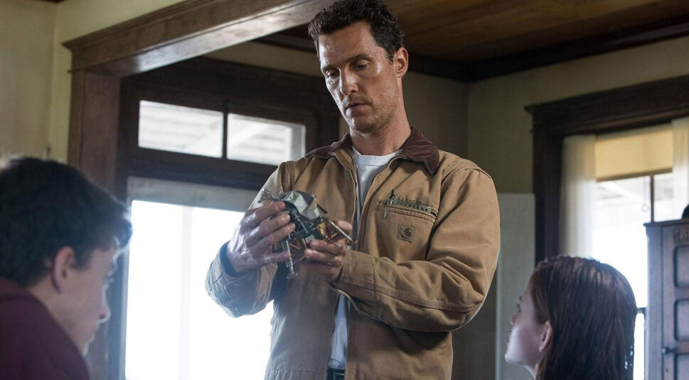 Auf diesem Bild zu Interstellar Fakten ist Cooper, gespielt von Matthew McConaughey, in einer nachdenklichen Szene zu sehen. Er hält ein Modell einer Raumsonde in den Händen und betrachtet es intensiv. Um ihn herum sitzen seine Kinder, die gespannt auf ihn blicken. Die häusliche Umgebung verstärkt den Kontrast zwischen Coopers Rolle als Vater und seiner zukünftigen Mission ins All, ein zentrales Thema des Films.