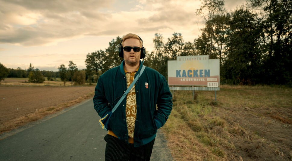 Auf dem Bild zur Serie Kacken an der Havel ist Anton "Fatoni" Schneider allein auf einer Landstraße zu sehen. Er trägt Kopfhörer, Sonnenbrille und eine auffällige Jacke, während er ernst in die Kamera blickt. Im Hintergrund steht ein Ortsschild mit der Aufschrift „Willkommen in Kacken an der Havel“. Die Stimmung wirkt ruhig und leicht ironisch.