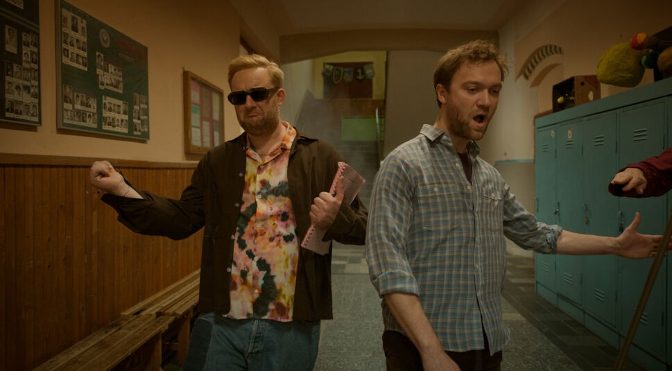 In einer Szene aus Kacken an der Havel gehen Anton „Fatoni“ Schneider und Vincent Redetzki durch einen Schulflur. Anton trägt Sonnenbrille und ein buntes Hemd, während er ein Notizbuch hält. Vincent schaut überrascht zur Seite. Die beiden wirken entschlossen und leicht überdreht, was den schrägen Humor von Kacken an der Havel unterstreicht.