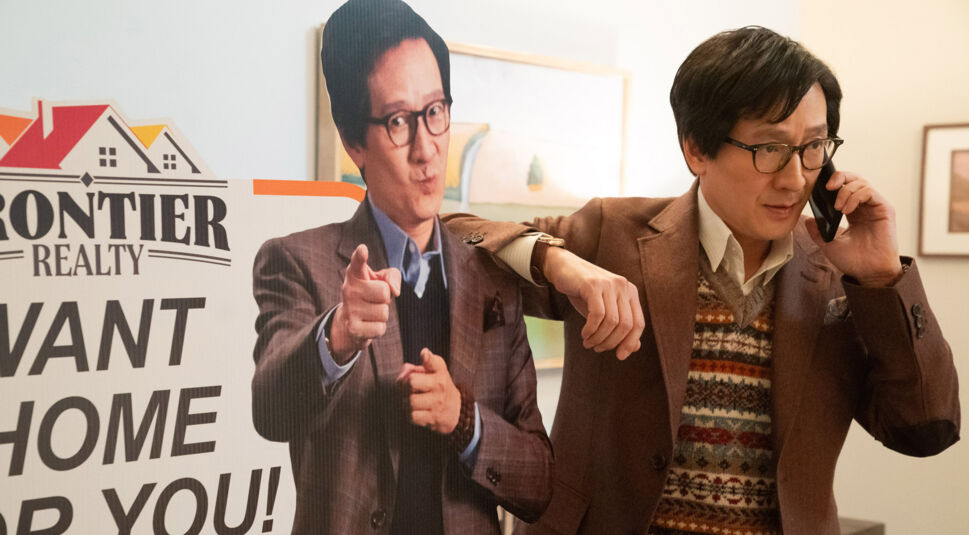 In einer Szene aus Love Hurts - Liebe tut weh steht Ke Huy Quan rechts und telefoniert konzentriert mit einem Handy. Neben ihm lehnt ein lebensgroßer Werbeaufsteller von ihm selbst, der mit einem Finger auf den Betrachter zeigt und den Schriftzug "Frontier Realty" trägt. Quan trägt ein braunes Sakko mit gemustertem Pullover, passend zur stilvollen Büroatmosphäre im Hintergrund.