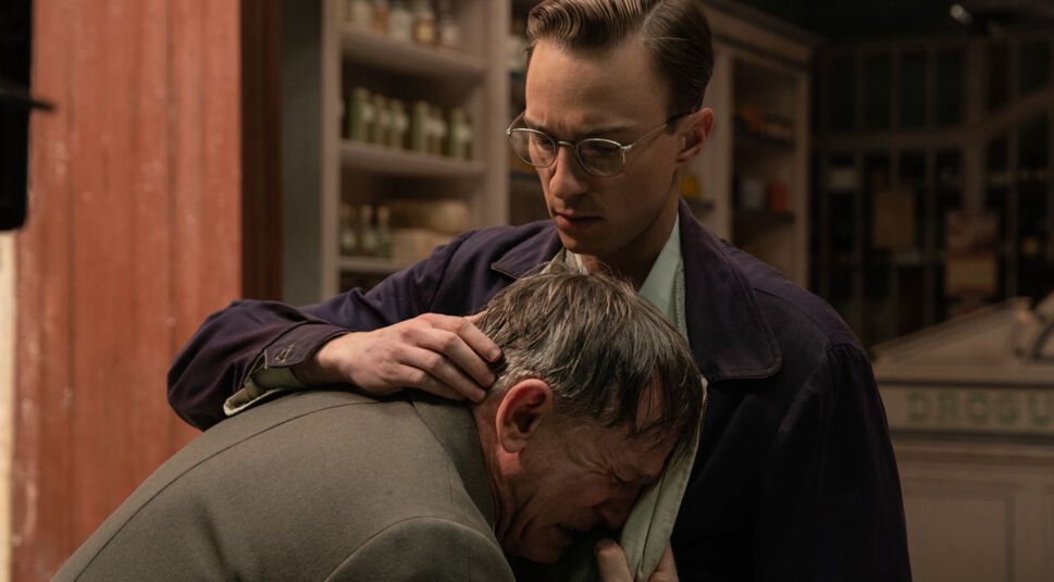 In dieser emotionalen Szene aus dem Film Queer umarmt Eugene Allerton, gespielt von Drew Starkey, den weinenden William Lee, dargestellt von Daniel Craig. Eugene hält William tröstend fest, während dieser sein Gesicht verzweifelt an Eugene lehnt. Die beiden befinden sich in einem Laden, erkennbar an den Regalen im Hintergrund. Die Szene drückt eine starke emotionale Verbundenheit und Unterstützung aus.