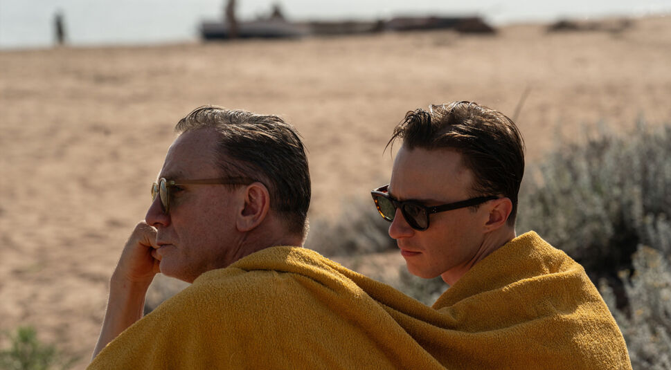 In dieser Szene aus dem Film Queer sitzen William Lee, gespielt von Daniel Craig, und Eugene Allerton, dargestellt von Drew Starkey, gemeinsam in ein gelbes Handtuch gehüllt am Strand. Beide tragen Sonnenbrillen und blicken nachdenklich in die Ferne, wobei die warme Strandumgebung eine ruhige und intime Atmosphäre schafft.