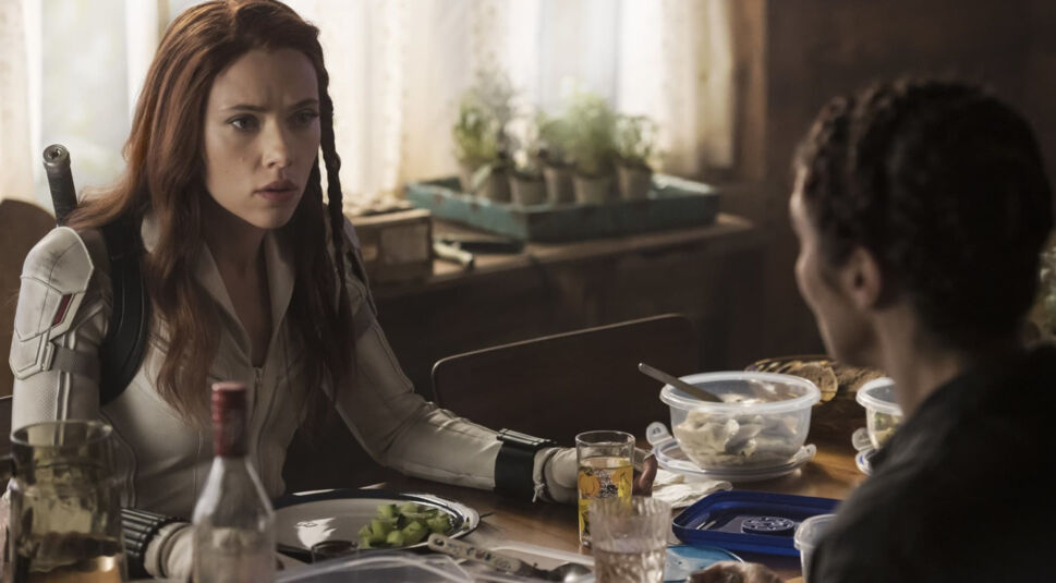 In einer Szene aus dem Marvel-Film Black Widow ist Scarlett Johansson als Natasha Romanoff alias Black Widow zu sehen. Sie trägt einen weißen Kampfanzug und sitzt an einem gedeckten Esstisch, während sie ernsthaft mit einer anderen Person spricht. Im Hintergrund sind Kräuterpflanzen und rustikale Einrichtungselemente zu erkennen.