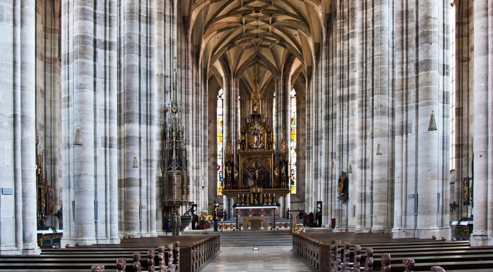 Das Bild zeigt das beeindruckende Innere der Kirche St. Georg in Dinkelsbühl, einen der Drehorte der Serie "Die Kaiserin". Die gotische Architektur wird durch die hohen Säulen, das kunstvolle Gewölbe und den reich verzierten Altar mit bunten Glasfenstern im Hintergrund hervorgehoben. Die Atmosphäre wirkt ehrwürdig und majestätisch.