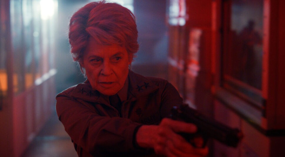 Auf dem Bild aus Stranger Things Staffel 5 ist Linda Hamilton als Dr. Kay zu sehen. Sie steht in einem rot beleuchteten Korridor, hält eine Pistole mit beiden Händen und blickt fokussiert und angespannt nach vorne. Ihre militärische Jacke und der entschlossene Gesichtsausdruck unterstreichen ihre kampferprobte und autoritäre Rolle.