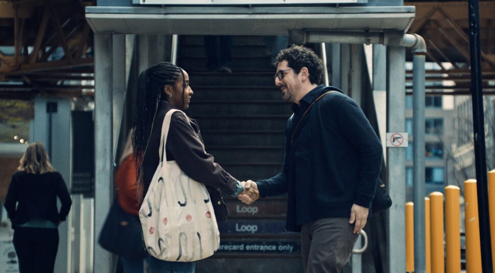 Auf dem Bild zur Serie The Bear Staffel 4 sind Sydney Adamu, gespielt von Ayo Edebiri, und Adam Shapiro, dargestellt von Adam Shapiro, zu sehen. Die beiden stehen vor einer Treppe im städtischen Bereich und schütteln sich die Hände. Sydney trägt eine große Stofftasche, während Adam eine Umhängetasche trägt. Die Szene wirkt alltäglich und zeigt eine Begegnung in einer urbanen Umgebung.