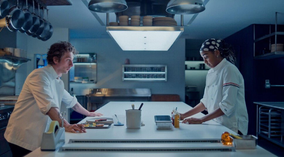 Auf dem Bild zur Serie The Bear Staffel 4 sind Carmen „Carmy“ Berzatto, gespielt von Jeremy Allen White, und Sydney Adamu, dargestellt von Ayo Edebiri, zu sehen. Beide stehen in einer modernen, professionellen Küche und wirken konzentriert. Carmy lehnt sich auf die Arbeitsfläche, während Sydney Zutaten vorbereitet. Die Szene ist in kühlem Licht gehalten und vermittelt eine angespannte, fokussierte Stimmung.