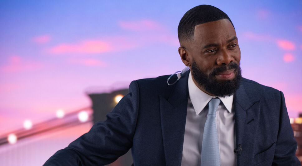Auf dem Bild zur Serie The Madness ist Colman Domingo als Muncie Daniels zu sehen. Er trägt einen eleganten dunkelblauen Anzug mit weißem Hemd und hellblauer Krawatte. Im Hintergrund erstreckt sich eine Brücke vor einem Himmel in Pastelltönen, der die Abendstimmung unterstreicht. Sein Gesichtsausdruck ist freundlich und gelassen, während ein Ohrhörer auf eine professionelle Szene hindeutet.