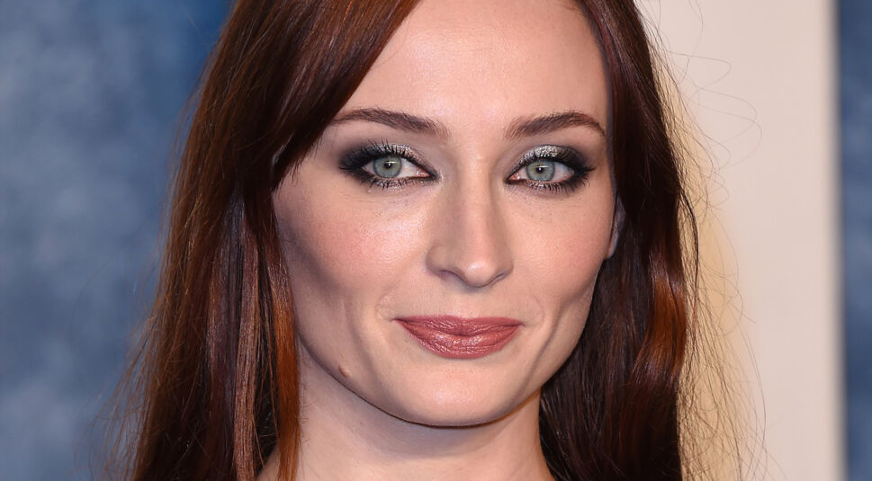 Auf dem Pressebild zur Tomb Raider-Serie auf Amazon Prime Video ist Schauspielerin Sophie Turner zu sehen, die im Gespräch für die Hauptrolle ist. Sie trägt ein dezentes Make-up mit betontem Lidschatten, ihr rötliches Haar fällt glatt über die Schultern. Der Hintergrund ist weich verschwommen und blau gehalten, was den Fokus auf ihr Gesicht lenkt.