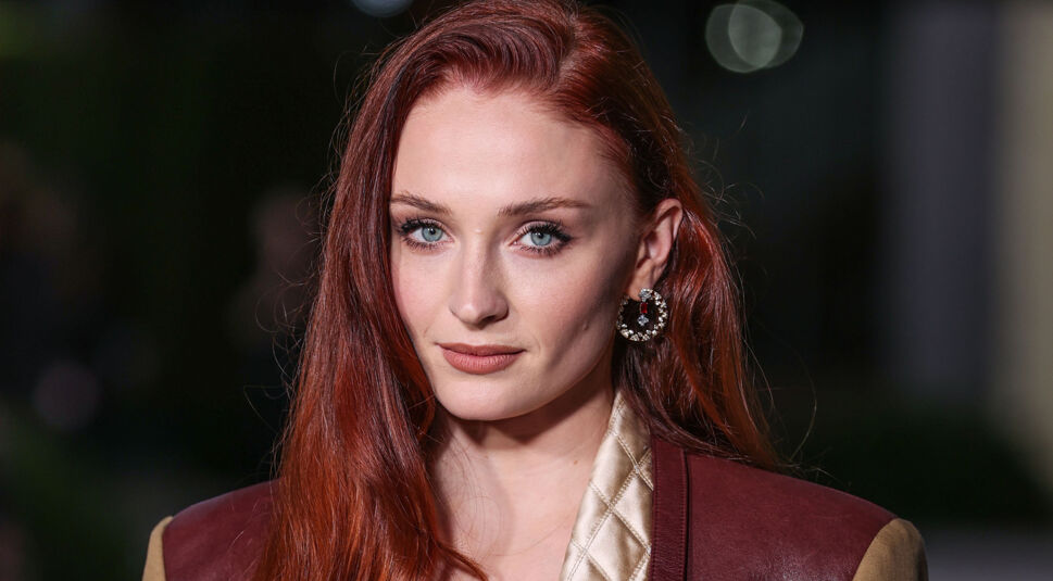 Auf dem Pressebild zur Tomb Raider-Serie auf Amazon Prime Video ist Sophie Turner zu sehen, die als mögliche Hauptdarstellerin gehandelt wird. Sie trägt ihr rotes Haar offen, dezentes Make-up und auffällige Ohrringe. Ihr Look wird durch eine gesteppte, beige Jacke mit braunen Lederakzenten ergänzt. Der Hintergrund ist dunkel mit weichen Lichtreflexen.