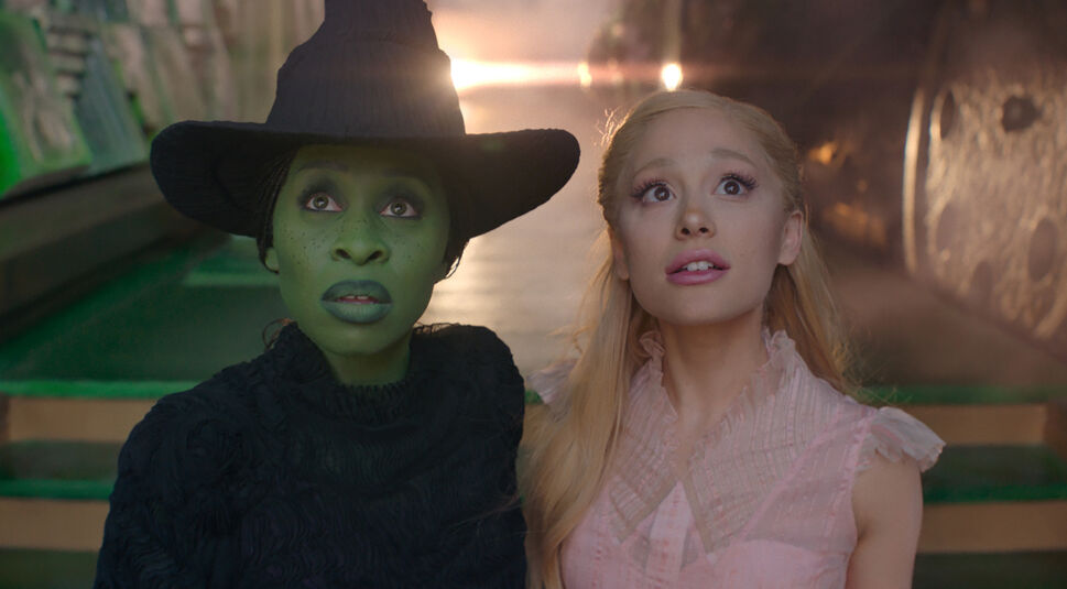 Auf dem Bild aus dem Wicked-Film sind Cynthia Erivo als Elphaba und Ariana Grande als Glinda zu sehen. Elphaba hat grüne Haut, trägt ein schwarzes Kleid und einen spitzen Hut, während Glinda in einem zarten rosa Kleid mit verträumtem Blick dargestellt ist. Beide schauen fasziniert in die Ferne, umgeben von einer mystischen und leuchtenden Atmosphäre.