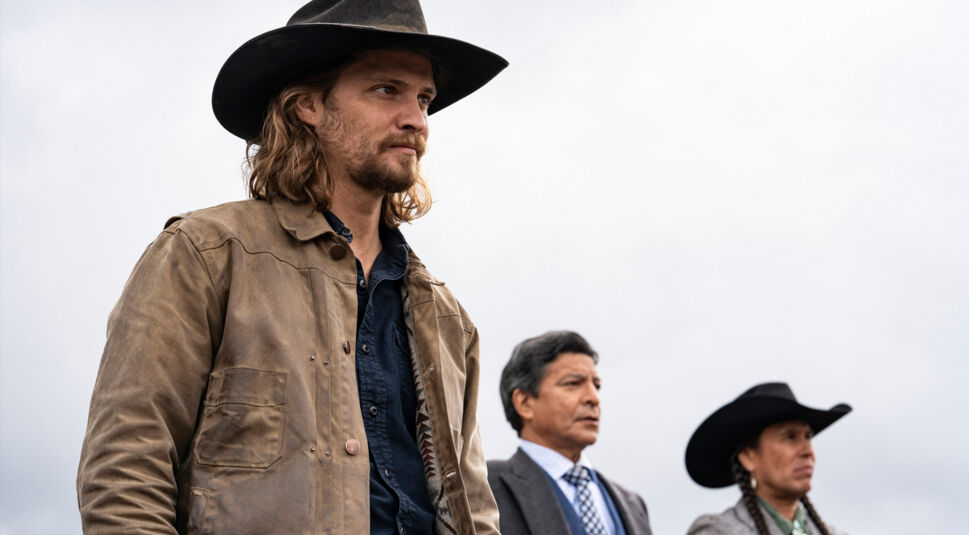 Auf dem Bild in dem Artikel über die Yellowstone Reihenfolge, sind drei Männer zu sehen. Ein Mann mit einem Cowboyhut steht ganz vorne, danach ein Mann in Anzug und dahinter ein weiterer Mann mit einem Cowboyhut. Alle drei schauen in die Ferne. Im Hintergrund sind dichte Wolken zu sehen.