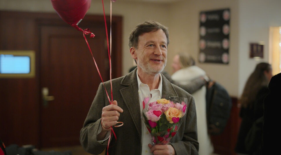 Auf dem Bild in dem Artikel über die Zeit Verbrechen-Serie, ist ein Mann zu sehen, der in der Mitte des Bildes steht und Blumen und Ballons in den Händen hält. Im Hintergrund sind eine Tür und weitere Menschen zu sehen.