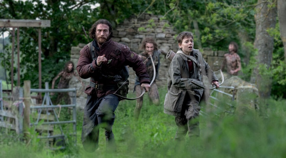 Auf dem Bild aus 28 Years Later sind Jamie, gespielt von Aaron Taylor-Johnson, und sein Sohn Spike, dargestellt von Alfie Williams, zu sehen. Beide laufen mit Bögen in der Hand vor Zombies weg.