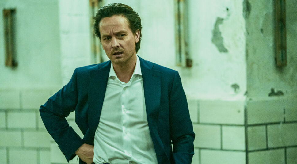 Auf dem Bild zu den besten Serien 2024 ist Tom Schilling als Björn Diemel in der Serie Achtsam Morden zu sehen. Er trägt einen dunkelblauen Anzug mit weißem Hemd und wirkt ernst und leicht erschöpft. Die Szenerie zeigt eine düstere, verlassene Umgebung mit kahlen, beschädigten Wänden und einem grünen Farbstich, was die spannungsgeladene Stimmung der Szene unterstreicht.