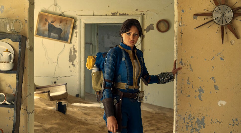 uf dem Bild zu den besten Serien 2024 ist Ella Purnell als Lucy in der Serie Fallout zu sehen. Sie trägt einen blauen Vault-Anzug mit gelben Details und ein futuristisches Gadget am Arm. Mit ernstem Blick und einem Rucksack steht sie in einem verfallenen Raum voller Sand. Die bröckelnden Wände, ein altes Bild und eine beschädigte Uhr verstärken die postapokalyptische Atmosphäre der Szene.