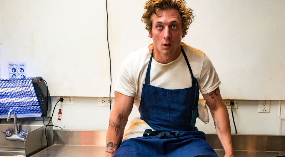 Auf dem Bild zu den besten Serien 2024 ist Jeremy Allen White in der Serie The Bear zu sehen. Er trägt eine weiße T-Shirt und eine blaue Kochschürze, sitzt in einer hektisch wirkenden Restaurantküche und wirkt erschöpft und konzentriert. Seine unordentlichen Haare unterstreichen die stressige Atmosphäre, die typisch für das Küchenumfeld der Serie ist.
