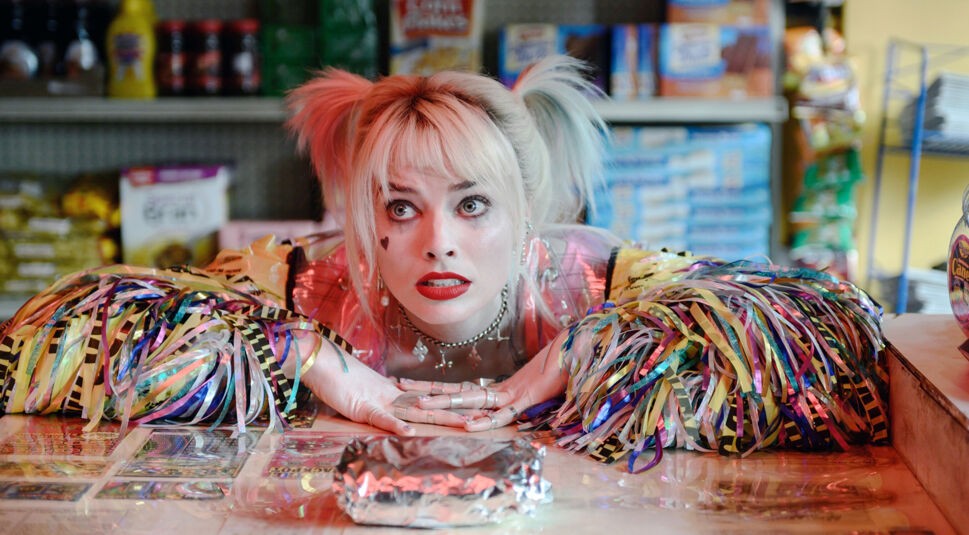 In dieser Szene aus dem DC Universum ist Harley Quinn, gespielt von Margot Robbie, zu sehen. Sie liegt mit neugieriger Miene und weit geöffneten Augen auf einem Verkaufstresen. Ihr auffälliges Outfit besteht aus bunten, schillernden Fransen und einer durchsichtigen Jacke. Im Hintergrund sind Regale mit Lebensmitteln und Produkten zu erkennen, was auf eine Szene in einem kleinen Laden hinweist.