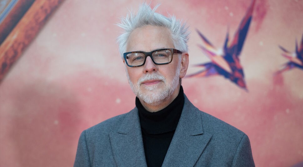 James Gunn, Regisseur und Drehbuchautor, ist auf diesem Bild zum DC Universum zu sehen. Er trägt eine graue Jacke über einem schwarzen Rollkragenpullover und schwarze Brillen. Sein markantes weißes Haar ist zerzaust. Der Hintergrund ist in Rosa- und Rottönen gehalten, mit stilisierten Raumschiff-Elementen, die auf eine futuristische oder filmische Kulisse hinweisen.