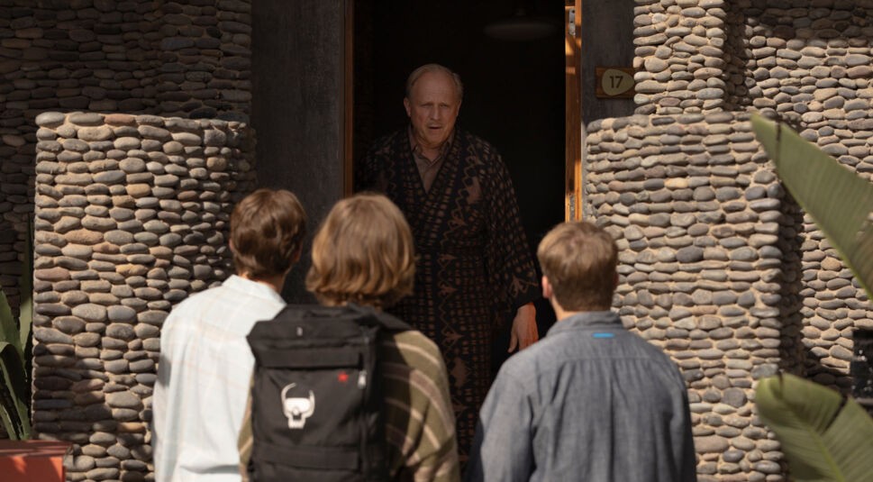 In einer Szene aus "Die drei ??? und der Karpatenhund" stehen Peter Shaw (Nevio Wendt), Bob Andrews (Levi Brandl) und Justus Jonas (Julius Weckauf) vor der Tür von Fenton Prentice (Ulrich Tukur). Fenton trägt einen gemusterten Bademantel und schaut die Jungen ernst an. Der Hintergrund zeigt eine Hauswand aus dekorativen Steinen.