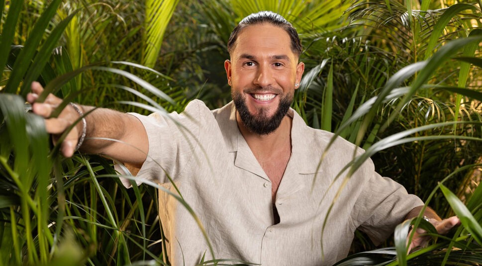 Auf dem Bild zum Dschungelcamp 2025 ist Maurice Dziwak zu sehen. Er trägt ein helles Leinenhemd und lächelt strahlend in die Kamera. Mit einer Hand greift er durch die dichten, grünen Palmenblätter, die für eine tropische Dschungelatmosphäre sorgen. Seine entspannte Haltung und das offene Lächeln vermitteln positive Energie.
