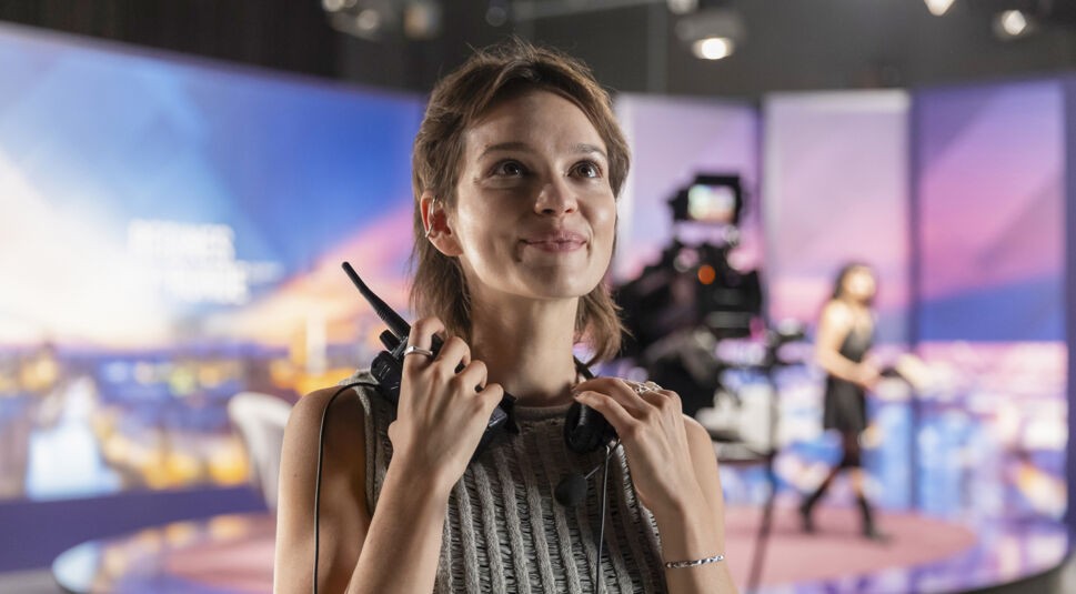 Auf dem Bild aus dem Film Wunderschön mit Karoline Herfurth ist Emilie Schüle in der Rolle der Julie zu sehen. Sie steht in einem TV-Studio und hält ein Headset sowie ein Walkie-Talkie in den Händen. Ihr Blick ist nachdenklich und leicht lächelnd nach oben gerichtet. Im Hintergrund sind verschwommen eine Kamerafrau und eine Studiokulisse mit violetten und blauen Farben zu erkennen. Die Szene vermittelt eine professionelle, aber inspirierende Atmosphäre.