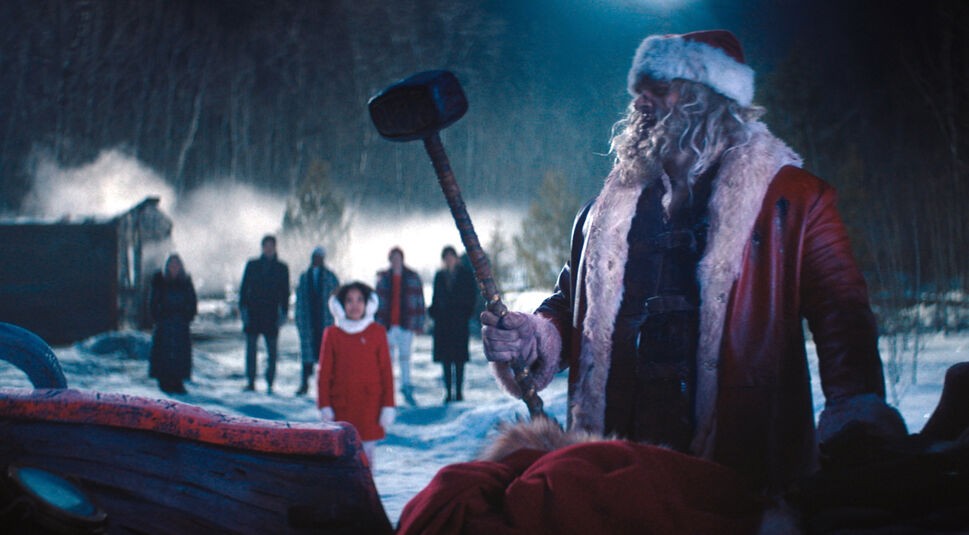 Auf dem Bild im GigaTV-Highlights Dezember 2024-Artikel siehst Du David Harbour als Santa in Violent Night. Santa steht in einer verschneiten, düsteren Landschaft und hält einen schweren Hammer entschlossen in der Hand. Im Hintergrund sind Leah Brady als Trudy und weitere Personen zu sehen, die gespannt die Szene verfolgen. 