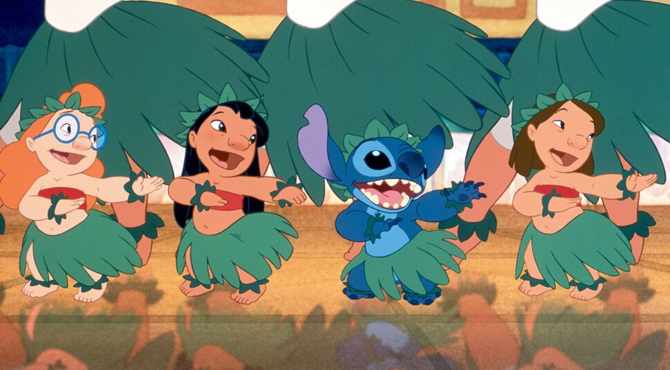 Auf dem Bild aus Lilo und Stitch tanzen Lilo, Stitch und zwei weitere Kinder in traditionellen hawaiianischen Hula-Outfits mit grünen Blätterröcken und Blattkränzen. Lilo, in der Mitte links, lächelt und bewegt die Arme rhythmisch, während Stitch, rechts neben ihr, mit breitem Grinsen begeistert mittanzt. Die beiden anderen Kinder strahlen ebenfalls Freude aus. Die Szene ist farbenfroh und vermittelt eine fröhliche, tropische Atmosphäre.