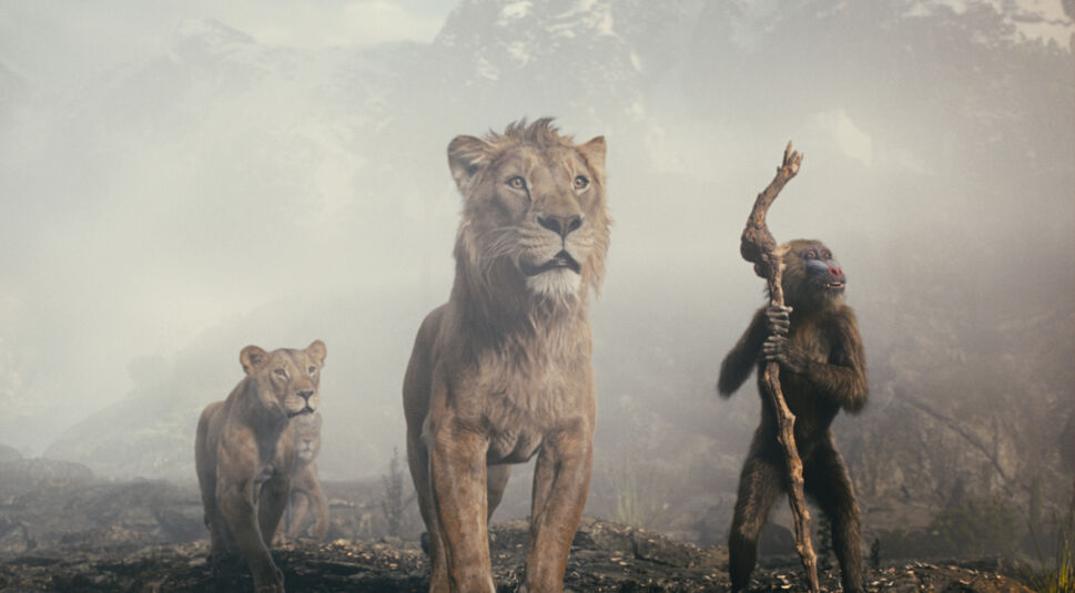 Auf dem Bild zur Mufasa Kritik sind drei ikonische Charaktere aus einer Szene des Films zu sehen. In der Mitte steht ein imposanter Löwe mit goldener Mähne und ernster Miene. Links neben ihm läuft eine Löwin mit fokussiertem Blick, während rechts ein Pavian mit einem Holzstab in der Hand steht und nachdenklich wirkt. Der Hintergrund zeigt eine nebelige, felsige Landschaft, die eine mystische Stimmung erzeugt.