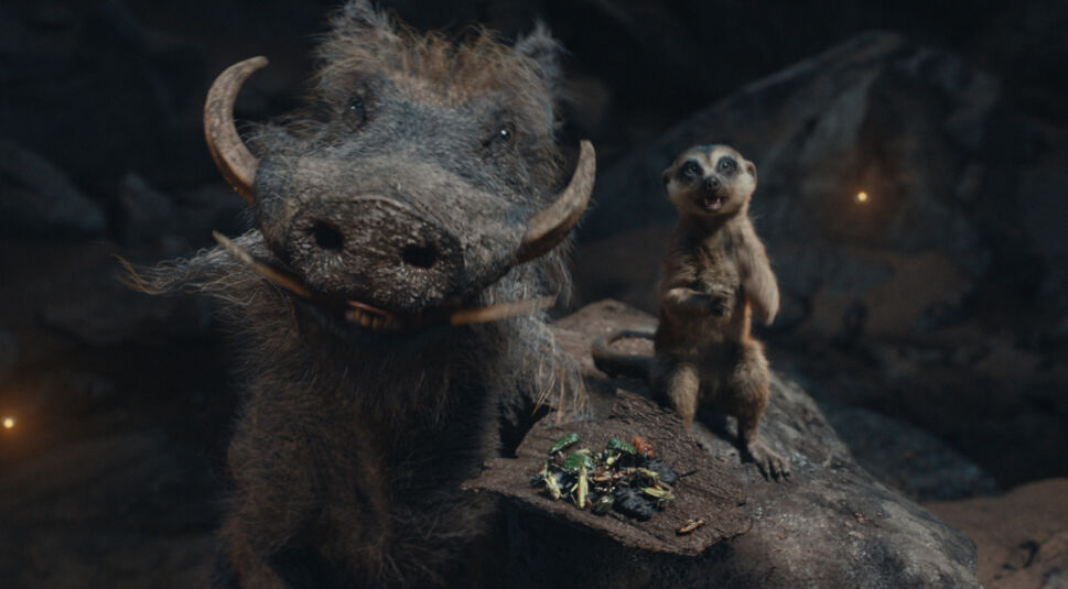 Auf dem Bild zur Mufasa Kritik sind Pumbaa, ein großes Warzenschwein mit imposanten Stoßzähnen, und Timon, ein kleines Erdmännchen, zu sehen. Pumbaa blickt freundlich in die Kamera, während Timon begeistert neben ihm steht und auf ein Holzbrett mit bunten Insekten deutet. Die Szene spielt in einer dunklen Höhle mit leichten Lichtreflexen im Hintergrund, was die lebendige und humorvolle Stimmung der beiden Charaktere unterstreicht.