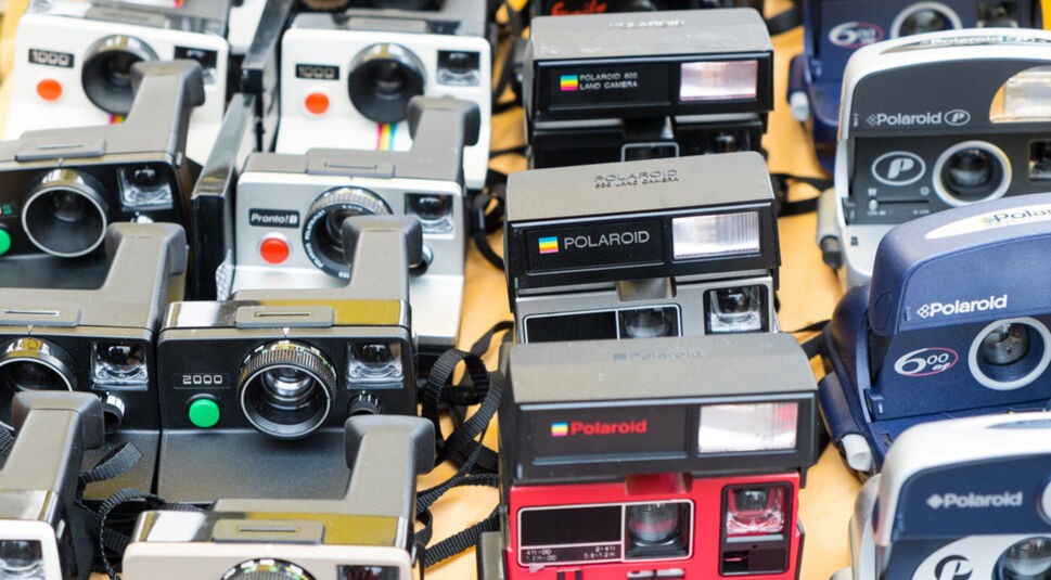 Auf dem Bild in dem Artikel über Retro-Gadgets, die ein Comeback machen sollten, ist eine Sammlung von Polaroid-Kameras in verschiedenen Modellen und Farben zu sehen. Die Kameras sind dicht nebeneinander aufgestellt, darunter klassische Modelle wie die Polaroid 1000 und Polaroid 2000. Einige Geräte haben schwarze, graue oder bunte Akzente, während das typische Retro-Design der 80er und 90er Jahre hervorsticht.